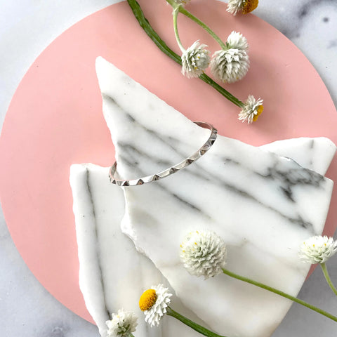 The SILVER CANAL BRACELET by Sarah Cecelia is displayed on white marble atop a pink circle, surrounded by delicate white and yellow flowers on a light background, highlighting elegant handmade silver jewelry.