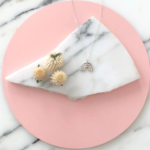 The Sarah Cecelia SILVER ARCO PENDANT and three cream-colored dried flowers sit atop a white marble slab, displayed on a round pink background over a marble surface.