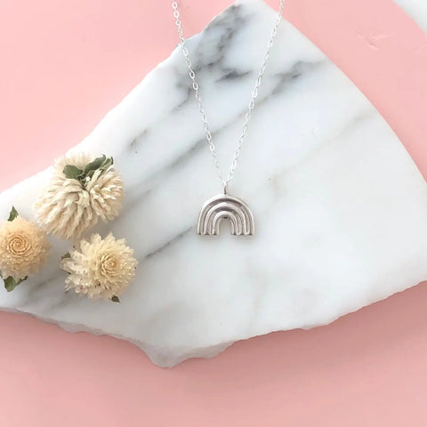 The Sarah Cecelia SILVER ARCO PENDANT, a handmade silver rainbow necklace, rests on a white marble slab with gray veins alongside three cream dried flowers, set against a pale pink background.