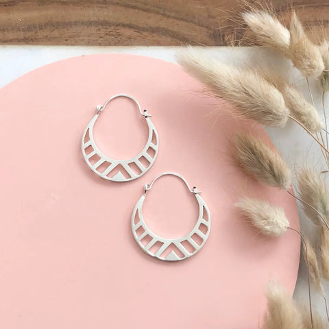 The SILVER SPECTRA HOOPS by Sarah Cecelia are handcrafted sterling silver earrings with geometric cutouts, shown on a round pink surface with dried pampas grass and a wooden table in the background.