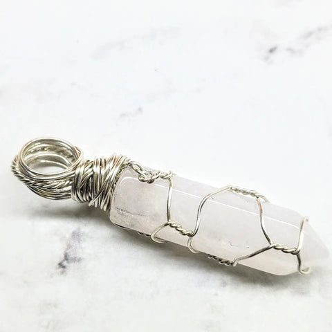 The Sterling Silver Messy Wire Wrap Gemstone Pointed Crystal Pendant by Alexa Martha Designs features clear quartz, intricately wrapped in sterling silver, and is displayed elegantly on a white marble surface.