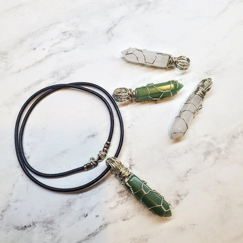 Four Alexa Martha Designs Sterling Silver Messy Wire Wrap Gemstone Pointed Crystal Pendants—two green, two white—are displayed on a white marble surface beside a black cord necklace with sterling silver clasps.