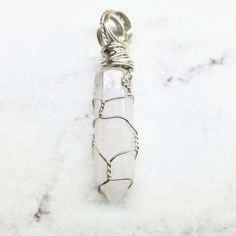 The Sterling Silver Messy Wire Wrap Gemstone Pointed Crystal Pendant by Alexa Martha Designs features a clear quartz crystal wrapped in sterling silver, showcased elegantly on a white marble surface.