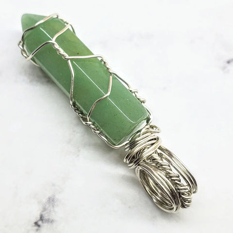 The Sterling Silver Messy Wire Wrap Gemstone Pointed Crystal Pendant by Alexa Martha Designs features an elegant green crystal wrapped in sterling silver, displayed on a white marbled surface with a loop for hanging.