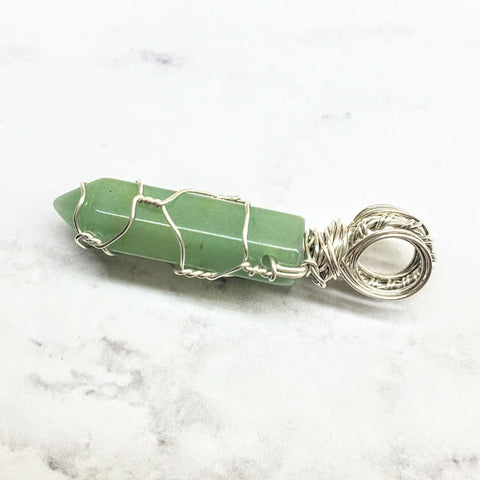 The Sterling Silver Messy Wire Wrap Gemstone Pointed Crystal Pendant by Alexa Martha Designs features a green gemstone in intricate silver wire with a decorative loop, elegantly displayed on a light marble surface.