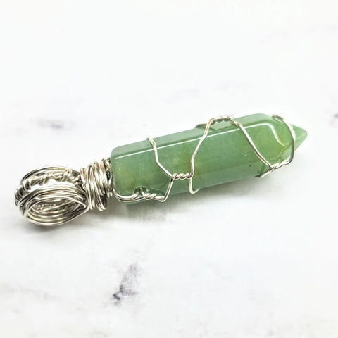 The Sterling Silver Messy Wire Wrap Gemstone Pointed Crystal Pendant by Alexa Martha Designs features a green gemstone in a decorative silver wire cage with a detailed loop, displayed on white marble.