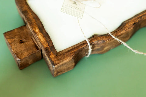 A close-up of the Shanti + Tula Handcrafted Trough 3-Wick Candle in a reusable wood bowl, featuring a rustic uneven edge and visible grain, with a tag tied by twine, resting on a light green surface. Made in the USA with soy wax.