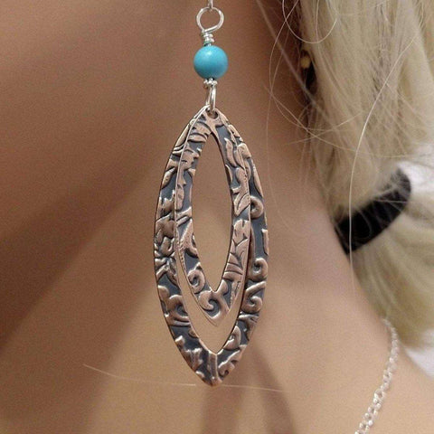 A close-up shows a person with light blonde hair wearing Alexa Martha Designs Turquoise Copper Embossed Pointed Oval Earrings featuring intricate floral patterns, a small turquoise bead, and sterling silver ear wires, along with a thin necklace.