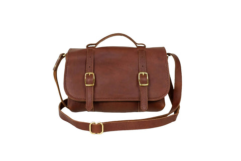 The Urban Southern Carriage Crossbody is a vintage-style brown leather satchel with a top handle, two front buckle straps, and a long adjustable shoulder strap—ideal as a leather crossbody bag on a white background.