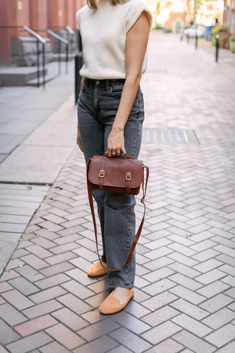 A woman stands on a paved walkway wearing a sleeveless cream top, gray jeans, tan flats, and holding the Urban Southern Carriage Crossbody handbag at her side. Her face is out of frame.