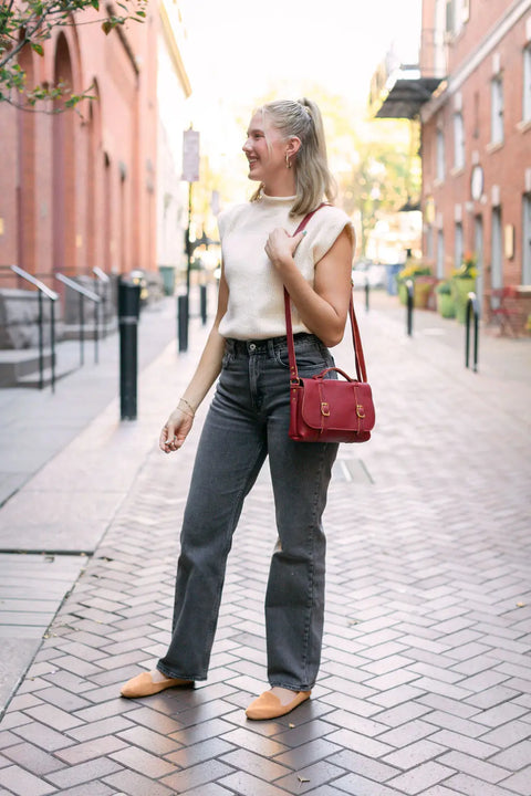 A blonde woman smiles on a brick-paved street, wearing a white sleeveless top, gray jeans, tan shoes, and carrying the Urban Southern Carriage Crossbody. Brick buildings and trees are visible in the background.
