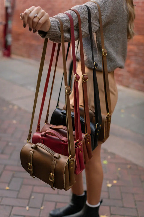 Outdoors against a brick wall, a person holds four Urban Southern Carriage Crossbody bags in brown, red, and black leather with gold hardware, highlighting their vintage style.