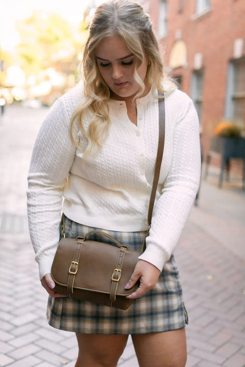 A woman with long blonde hair in a white textured sweater and plaid skirt carries the Urban Southern Carriage Crossbody while walking on a brick-paved street lined with buildings.
