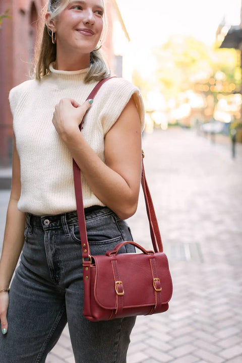 A woman in a sleeveless cream sweater and high-waisted gray jeans stands on a brick street, smiling softly. She carries the Urban Southern Carriage Crossbody over her shoulder. The background is gently blurred.