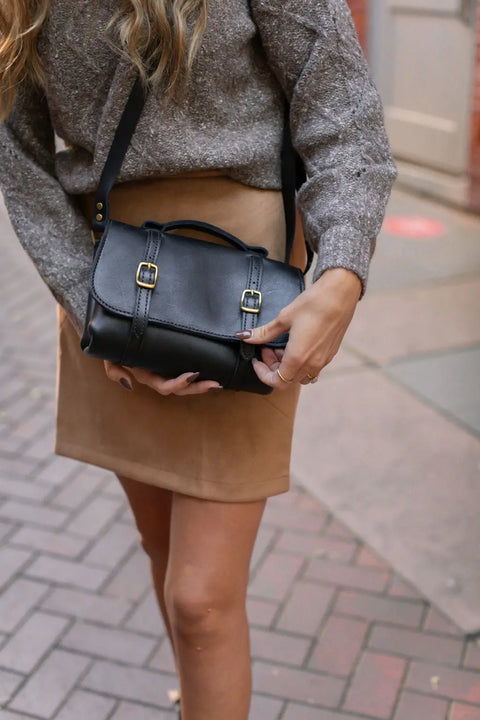 A woman in a gray sweater and tan skirt holds the Urban Southern Carriage Crossbody, a vintage-style handbag with gold buckles, while standing on a brick sidewalk.