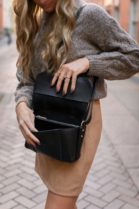 A woman with long wavy hair wears a brown sweater and tan skirt, standing on a cobblestone street as she opens an Urban Southern Carriage Crossbody, a vintage-style handbag handcrafted in the USA, to look inside.