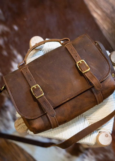 The Urban Southern Carriage Crossbody, handcrafted in the USA with rich brown leather and brass buckles, rests on a woven stool. Its vintage style echoes classic charm, with a cowhide rug partially visible in the background.
