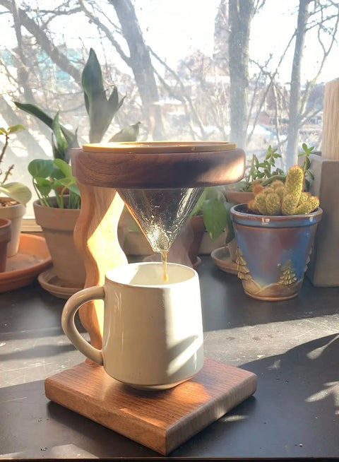 The Squiggle Pour Over 2 by Shop Cat Woodworks brews coffee as it drips into a ceramic mug. Sunlight filters through a window, highlighting potted plants and casting gentle shadows across the table.