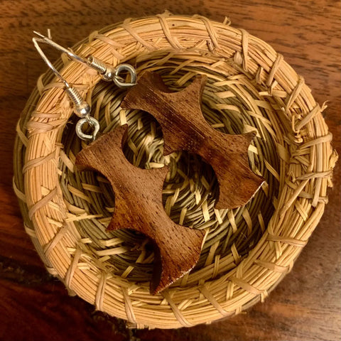 The Thorn Earring by Shop Cat Woodworks, handcrafted from walnut wood and shaped like apple cores, is displayed in a small round woven straw basket on a wooden surface.