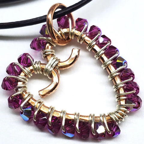 The Wire Wrapped Open Heart Crystal Necklace by Alexa Martha Designs showcases a heart-shaped pendant crafted from golden and silver wire, accented with purple crystal beads and a metallic Pisces symbol, all suspended from a black cord.