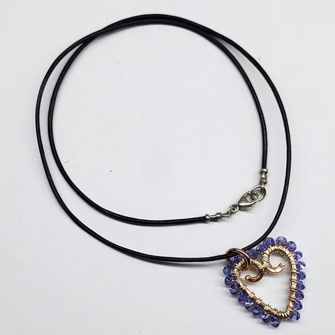 The Wire Wrapped Open Heart Crystal Necklace by Alexa Martha Designs features a handmade heart-shaped pendant wrapped in gold and copper wire with purple beads, displayed on a black cord—an elegant, unique accessory for any outfit.