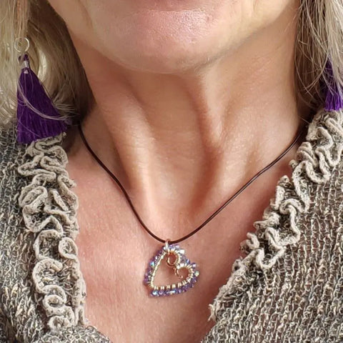 A woman wears the Alexa Martha Designs Wire Wrapped Open Heart Crystal Necklace, featuring a handmade purple heart pendant on a black cord, paired with a purple tassel earring and a textured beige top.
