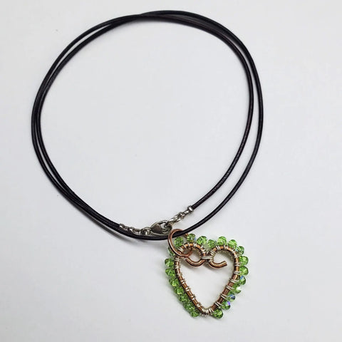 The Wire Wrapped Open Heart Crystal Necklace by Alexa Martha Designs features a handmade heart pendant with green beads, decorative copper wire swirls, and a black cord—an artistic and charming accessory.
