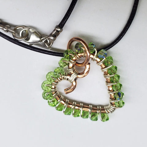 The Wire Wrapped Open Heart Crystal Necklace by Alexa Martha Designs features a heart-shaped pendant with silver wire and green beads on a black cord, adding artisanal charm to any outfit.