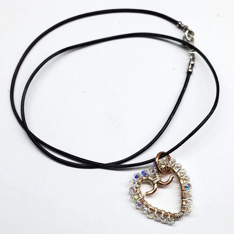 The Wire Wrapped Open Heart Crystal Necklace by Alexa Martha Designs features a handmade, heart-shaped pendant with clear faceted beads and a metal bow, beautifully displayed on a white background.