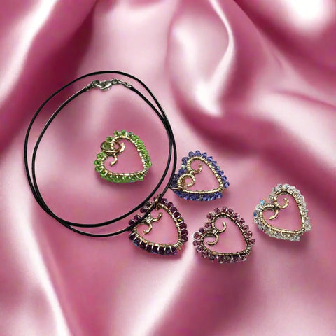Heart-shaped pendants in green, purple, pink, and clear crystals rest on pink satin; one hangs from a black cord, showcasing the elegance of the Alexa Martha Designs Wire Wrapped Open Heart Crystal Necklace.