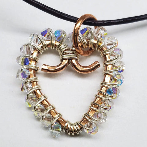The Wire Wrapped Open Heart Crystal Necklace by Alexa Martha Designs features a gold wire heart pendant with clear faceted beads and a central swirl, displayed on a black cord against a white background.