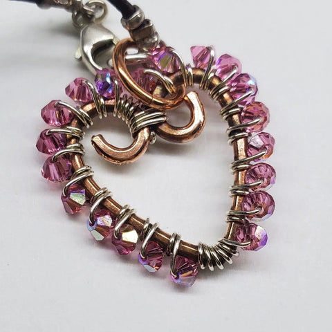 The Wire Wrapped Open Heart Crystal Necklace by Alexa Martha Designs features a pink crystal pendant with copper-toned spirals on a black cord, elegantly displayed against a white background.