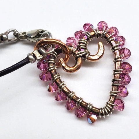 The Wire Wrapped Open Heart Crystal Necklace by Alexa Martha Designs features a handmade pendant with pink beads on a black cord and metal clasp, beautifully displayed against a white background.