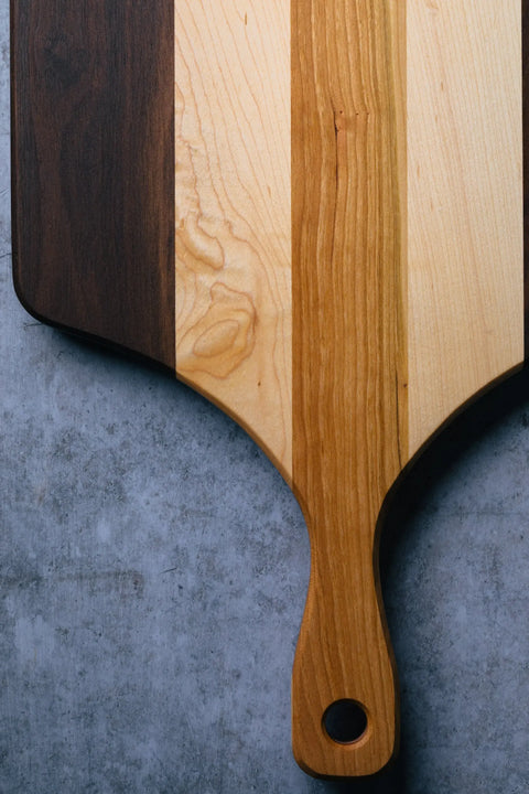 A close-up of The Bread Board - Standard by Montana Block, an American-made cutting board with a handle and three vertical wood tones, shown resting on a textured gray surface.
