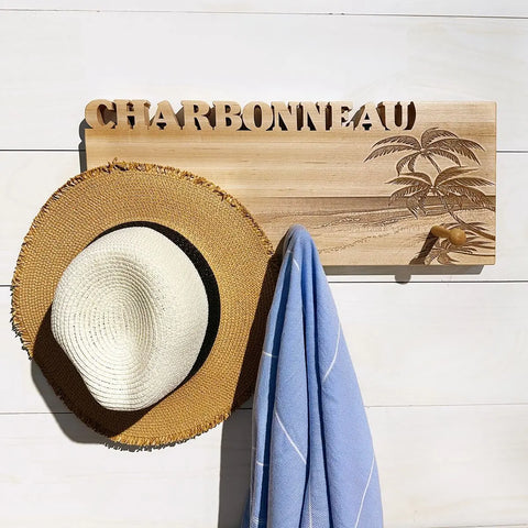 A straw hat and blue towel hang on the Handcrafted Wooden Coat Rack by Words with Boards, made in the USA, laser engraved with CHARBONNEAU and palm trees on a beach.