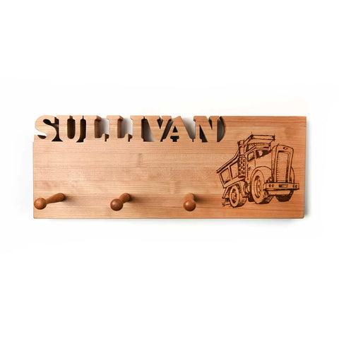 The Handcrafted Wooden Coat Rack by Words with Boards features three wood pegs, the name “SULLIVAN” cut out at the top, and a laser-engraved dump truck illustration on the right side. Made in the USA.