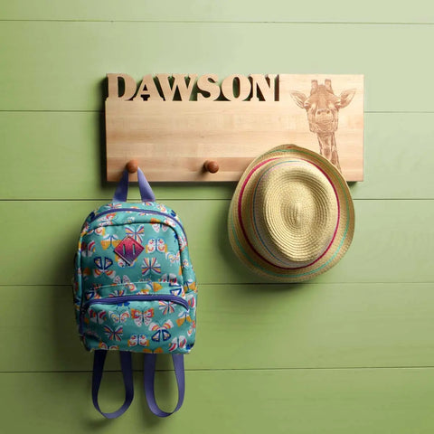 A Handcrafted Wooden Coat Rack by Words with Boards, featuring the name DAWSON and a giraffe design, displays a colorful straw hat and patterned blue backpack against a green wooden wall. Made in the USA.