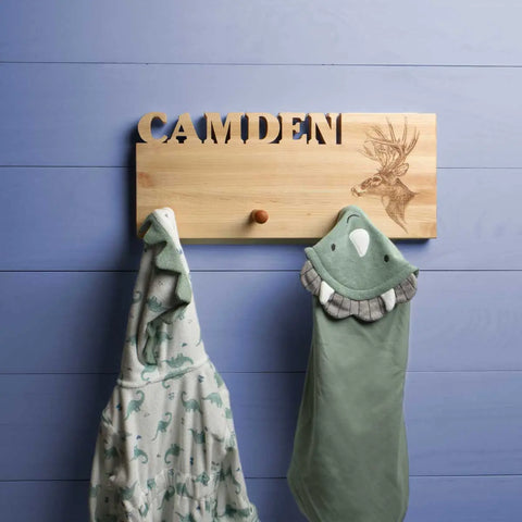 A Handcrafted Wooden Coat Rack made in the USA by Words with Boards features wood pegs and a stag engraving, holding two childrens hooded towels—one dinosaur print and one green bird—against a light blue wooden wall.