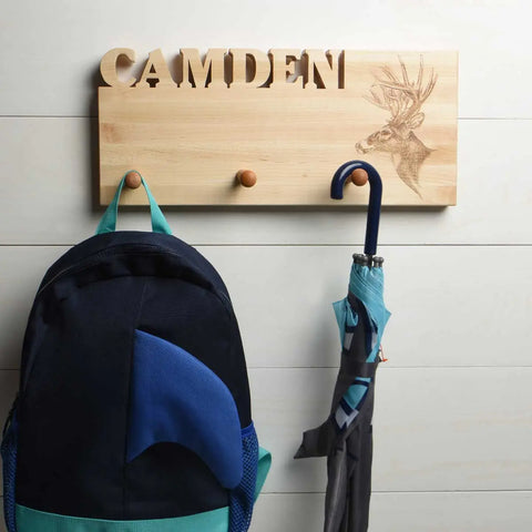The Handcrafted Wooden Coat Rack by Words with Boards features “CAMDEN” and a deer motif. A black and blue backpack hangs on one peg, with a closed umbrella on another, set against white wood paneling. Made in the USA.