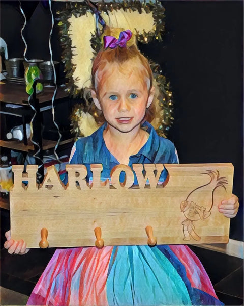 A young girl with a pink bow smiles, holding a Handcrafted Wooden Coat Rack by Words with Boards, laser engraved with HARLOW and wood pegs, featuring a cartoon troll in a colorful dress, surrounded by cheerful decorations.