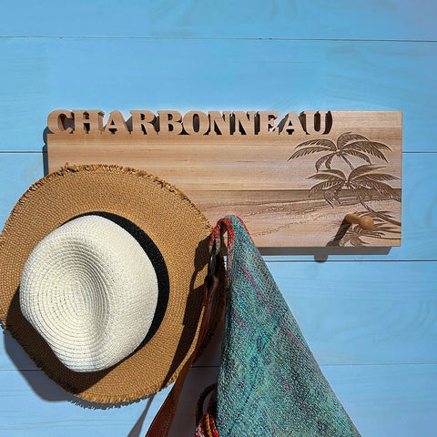 The Handcrafted Wooden Coat Rack by Words with Boards features CHARBONNEAU cut out at the top, a carved palm tree design, and wood pegs holding a straw hat and colorful bag, set against a light blue wooden wall. Made in the USA.
