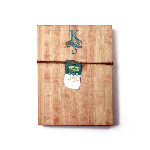 A handcrafted wooden cutting board by Words with Boards featuring a stone-inlay monogram, tied with a brown string and green tag. Photographed on a white background. Made in the USA.