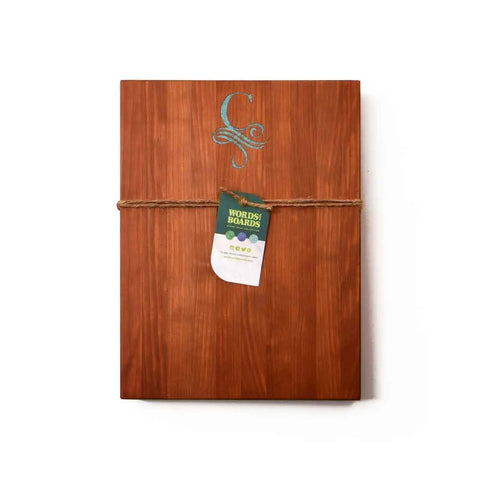A handcrafted wooden cutting board by Words with Boards, featuring a monogram with stone inlay, tied with twine and a green Words with Boards tag—made in the USA. Perfect personalized gift for any occasion. Background is white.