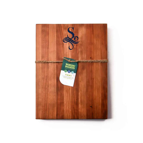 A handcrafted wooden cutting board by Words with Boards features a monogram with stone inlay, tied with twine and a green Words! Boards! tag—made in the USA. Makes a charming personalized gift. Shown on a white background.