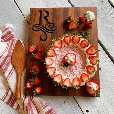 A pink strawberry pie with whipped cream and strawberries rests on the Handcrafted Wooden Cutting Board – Single Initial Monogram by Words with Boards, surrounded by fresh strawberries, a pie server, and a striped towel.