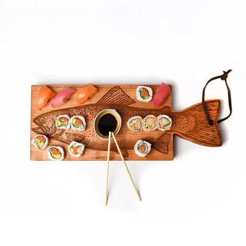 The Handcrafted Wooden Fish Shaped Cutting Board by Words with Boards (Made in USA) features a carved fish design, leather loop handle, and is perfect for serving sushi with its central soy sauce dish and chopstick rest.
