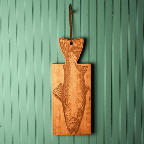 The Handcrafted Wooden Fish Shaped Cutting Board by Words with Boards adds unique kitchen flair as it hangs from a leather loop on a teal paneled wall—perfect for displaying artisanal, USA-made kitchenware.