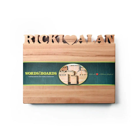 The Handcrafted Wooden Personalized Cutting Board (Small) by Words with Boards features “RICKI ♥ ALAN” cut out along the top and comes with a green Words with Boards branded band. Made in the USA.