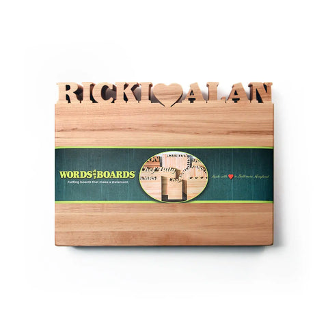The Handcrafted Wooden Personalized Cutting Board (Small) by Words with Boards features “RICKI ♥ ALAN” cut out along the top and comes with a green Words with Boards branded band. Made in the USA.