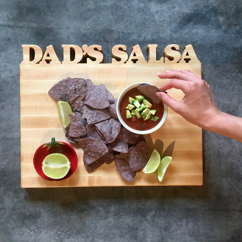 A hand dips a blue corn chip into salsa with avocado on a Words with Boards Handcrafted Wooden Personalized Cutting Board | Small—an ideal kitchen accessory and gift, crafted in the USA, shown with chips and lime wedges.